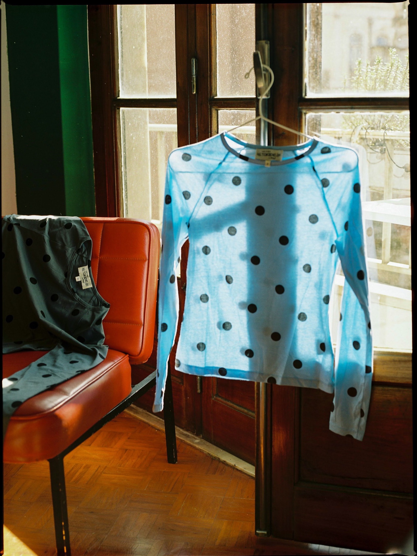 A blue polka-dot top on a hanger by a sunlit window, a dark polka-dot top over a red leather chair