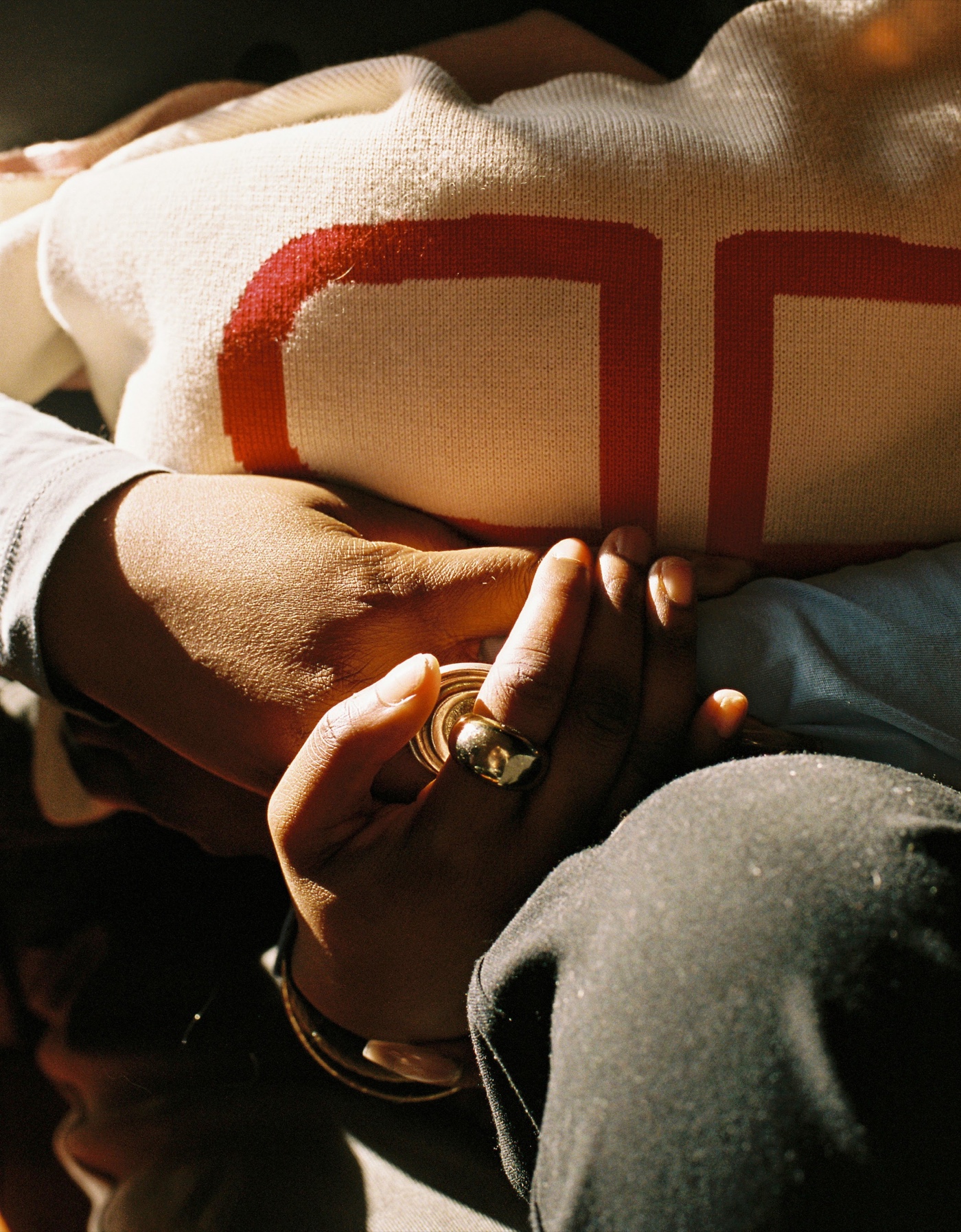 Crossed arms in a cream knit with a red motif, hands clasped, a gold ring catching the light
