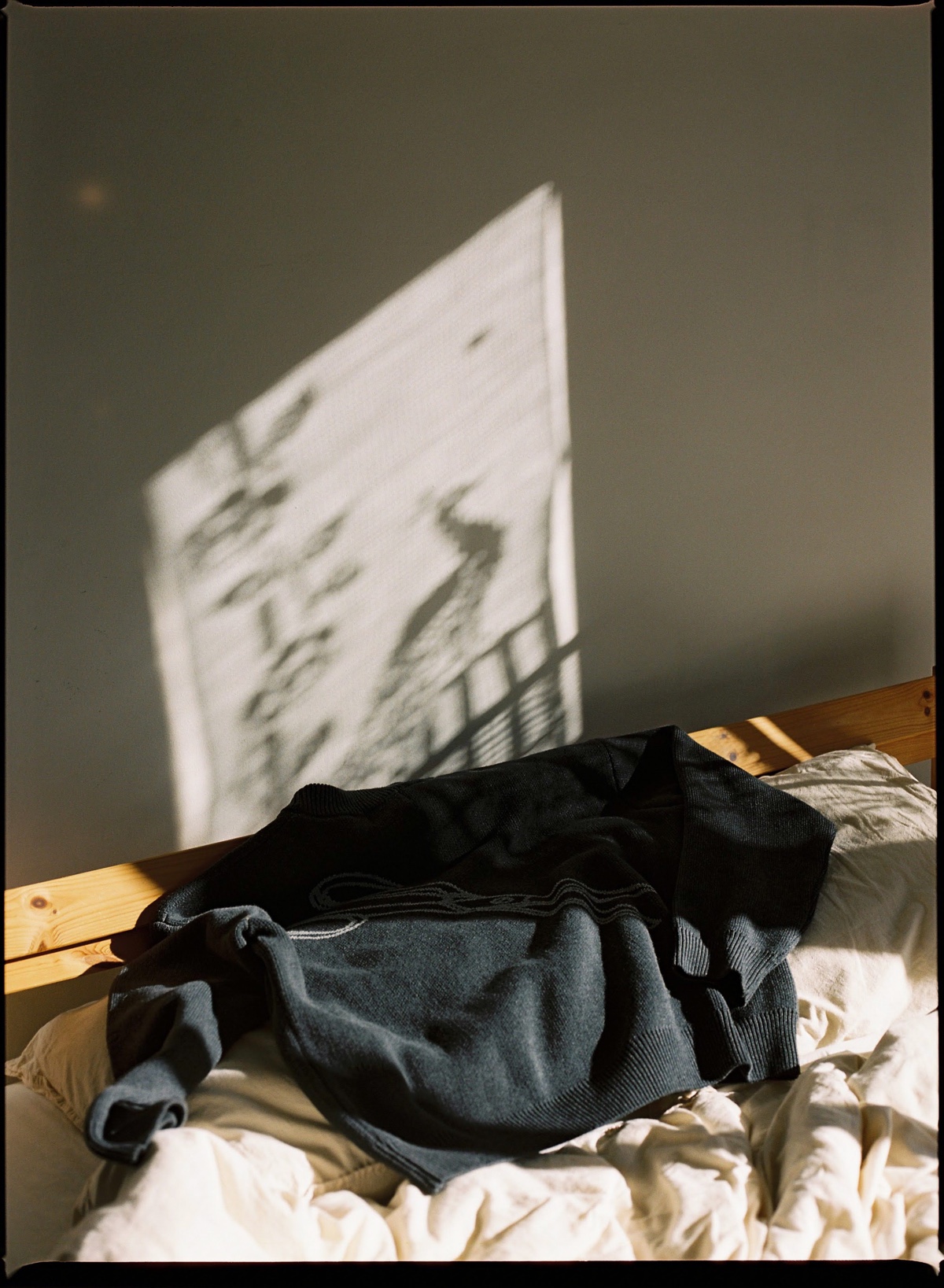 A dark knit resting on a sunlit bed, the window's shadow cast across the wall
