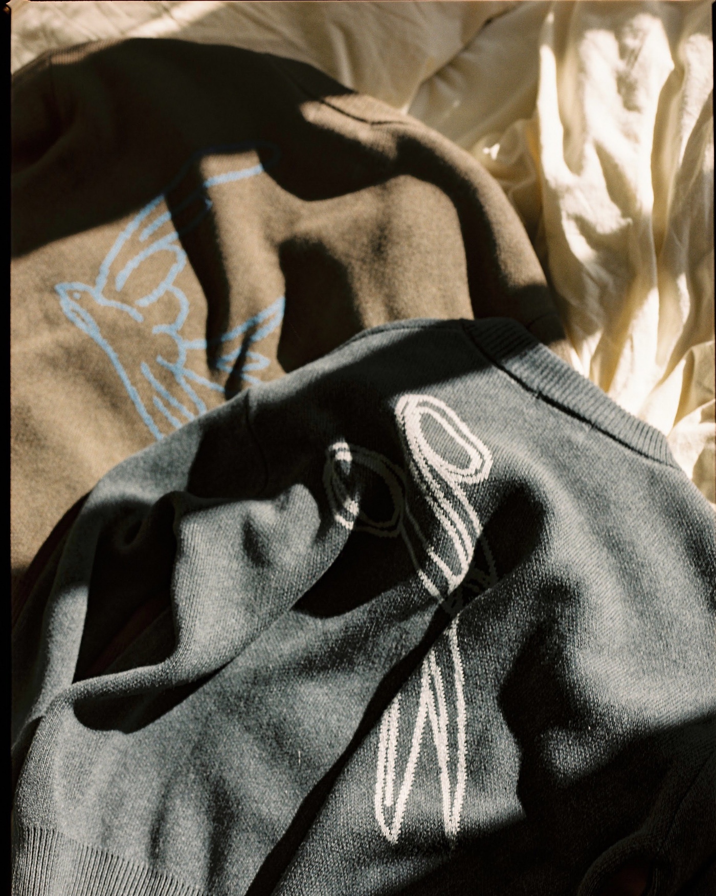 A tan knit with a blue bird motif and a grey logo knit resting in raking afternoon light