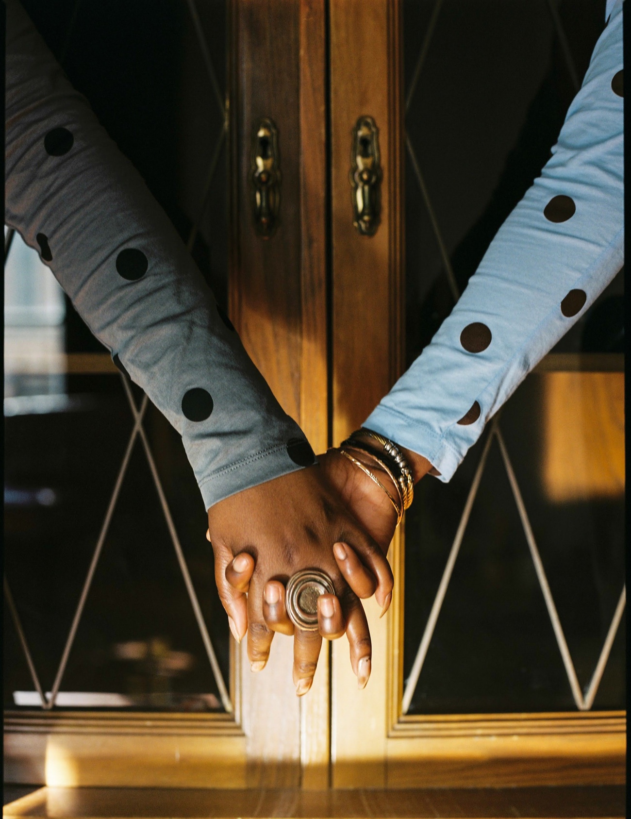 Two hands clasped over a wooden cabinet, polka-dot sleeves crossing, a gold ring catching the light
