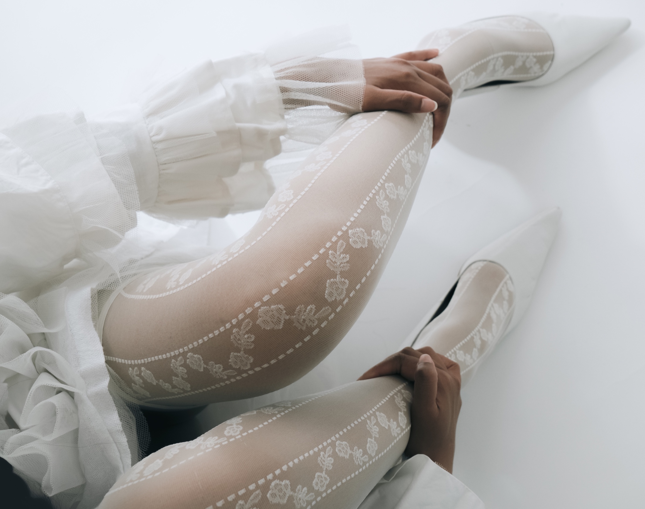 Legs in white floral lace tights and pointed heels, hands resting, against white