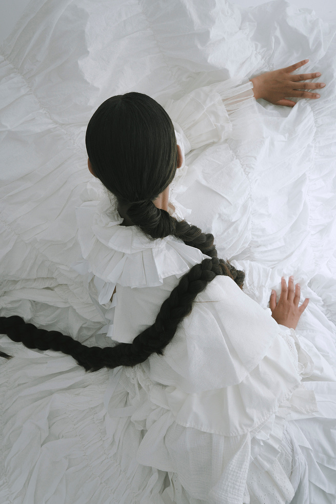 A figure with a long braid lying among white ruffled textiles, hands outstretched