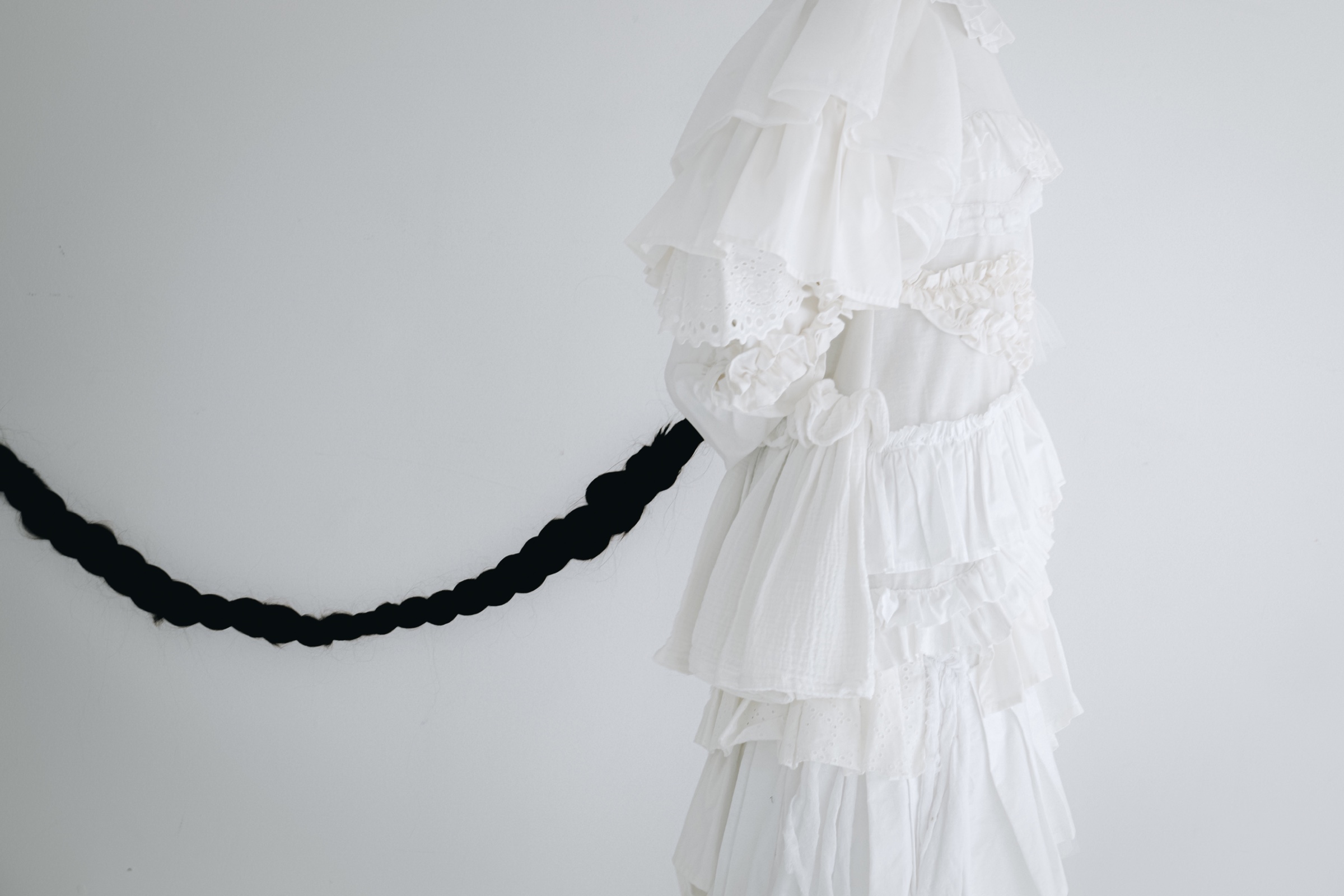 A white ruffled dress on a pale wall, a long black braid extending across to the left